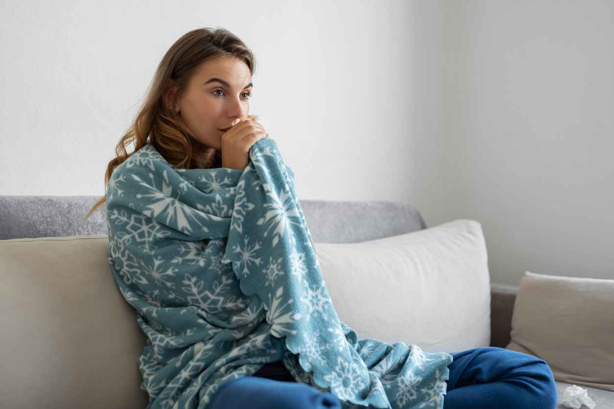 Woman sitting on a couch at home wrapped in a blanket due to a broken heater